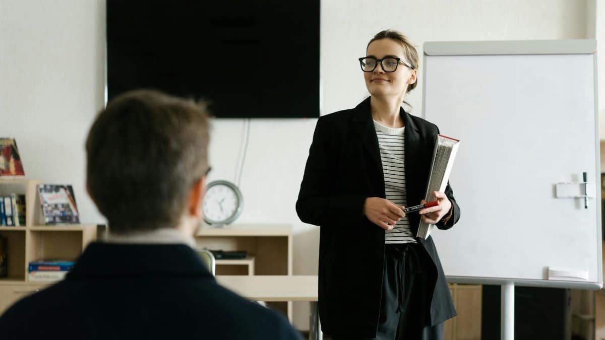 Caucasian female teacher in black blazer holding books, presenting to a student in a classroom.