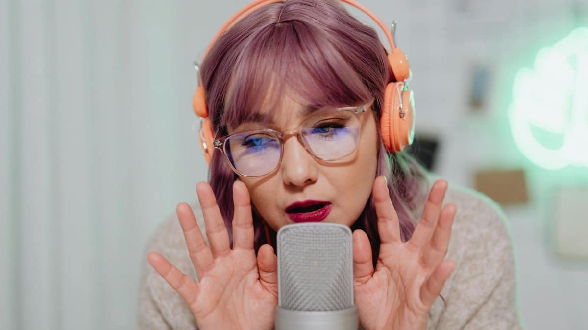 A woman with pink hair and headphones recording ASMR using a microphone indoors.