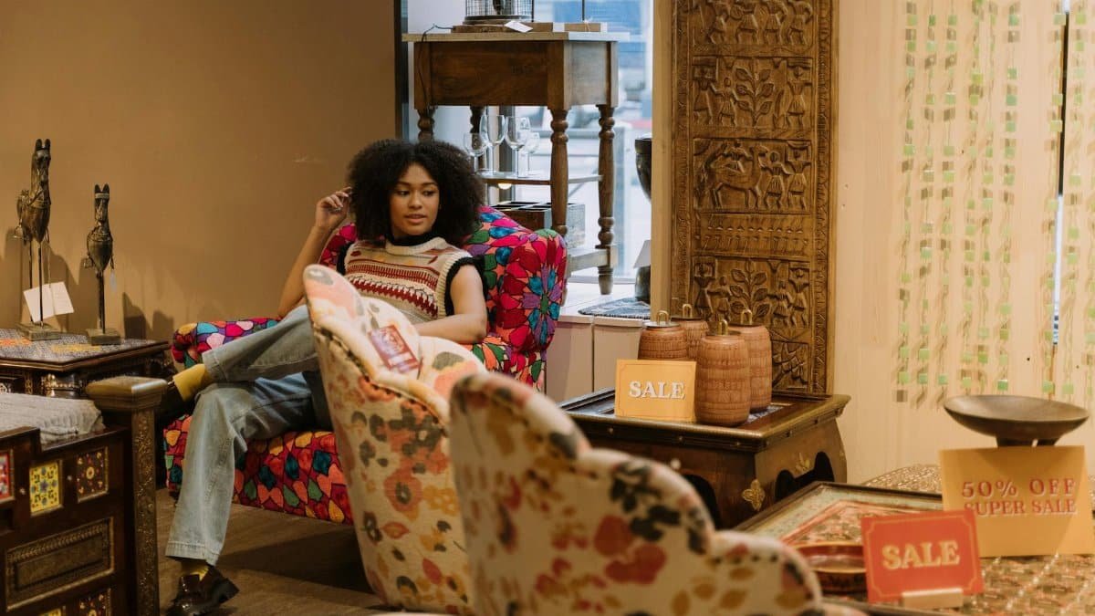 Woman with afro hair relaxing on a colorful chair in a furniture store showcasing sale signs and unique decor pieces.