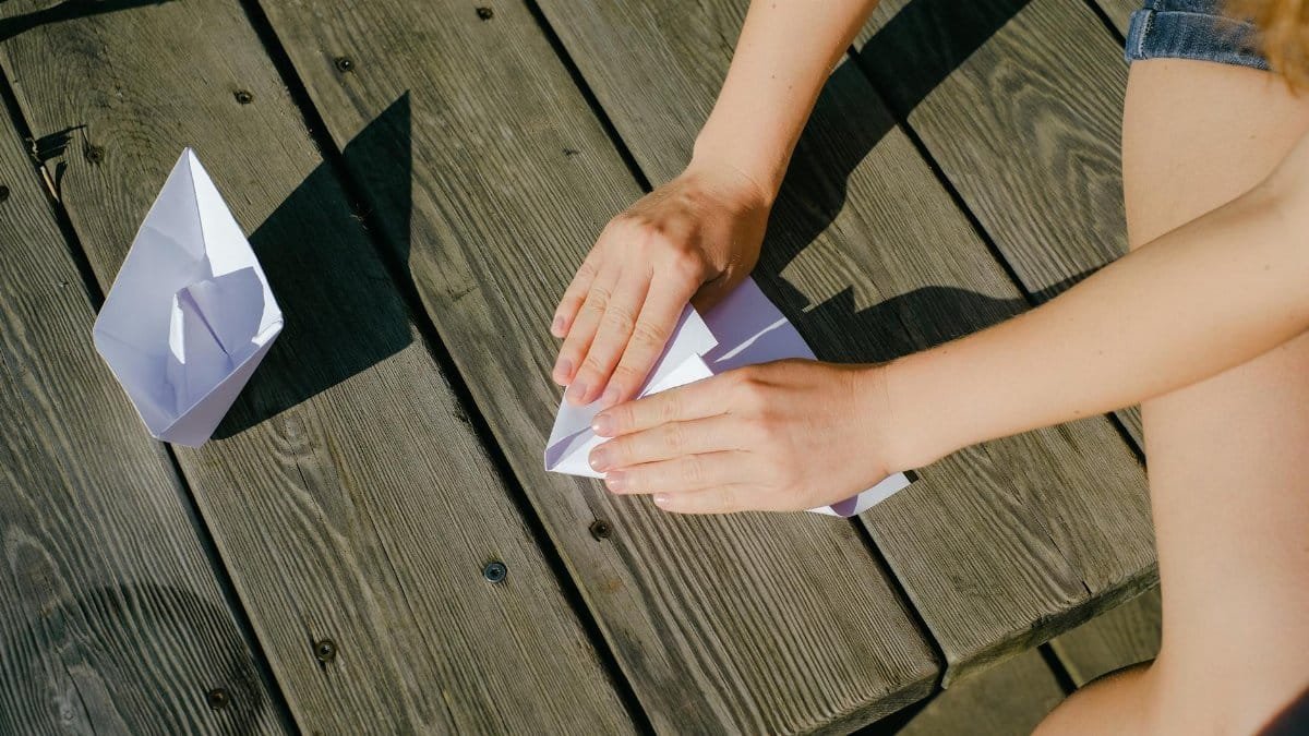 Hands folding paper boats on a wooden deck under the sun. Creative and peaceful outdoor activity.