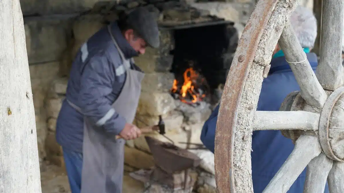 Blacksmith forging metal beside fire in rustic workshop, creating artisanal crafts.