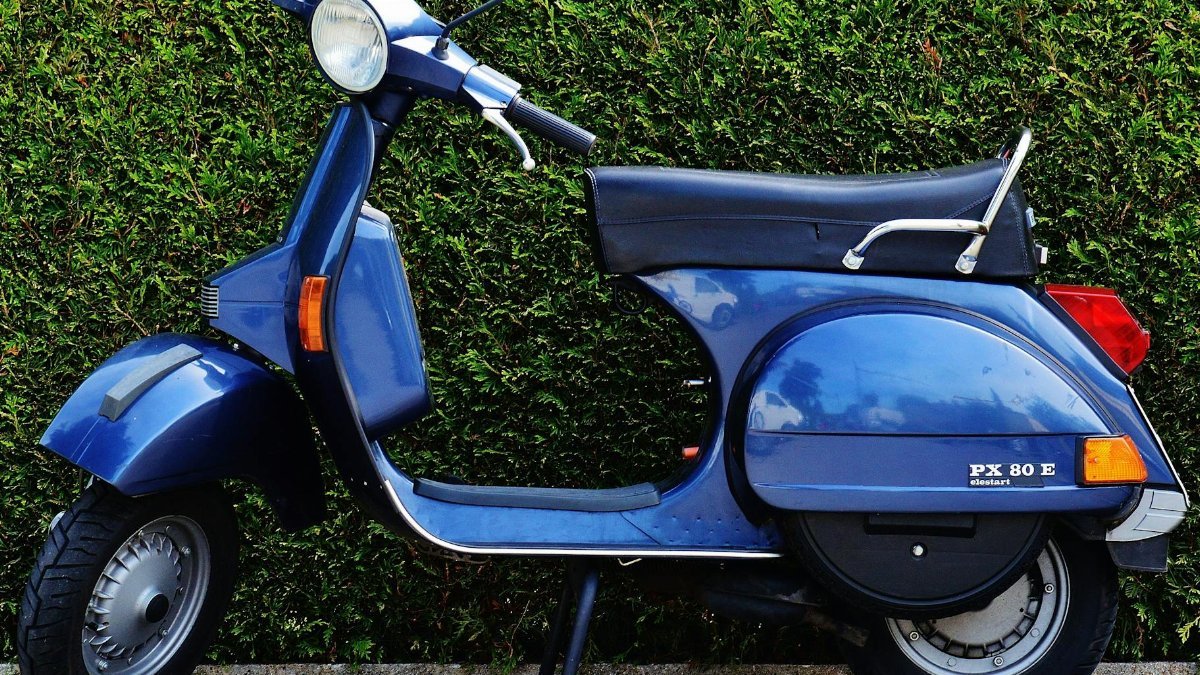 Vintage blue scooter parked by a green hedge, side view.