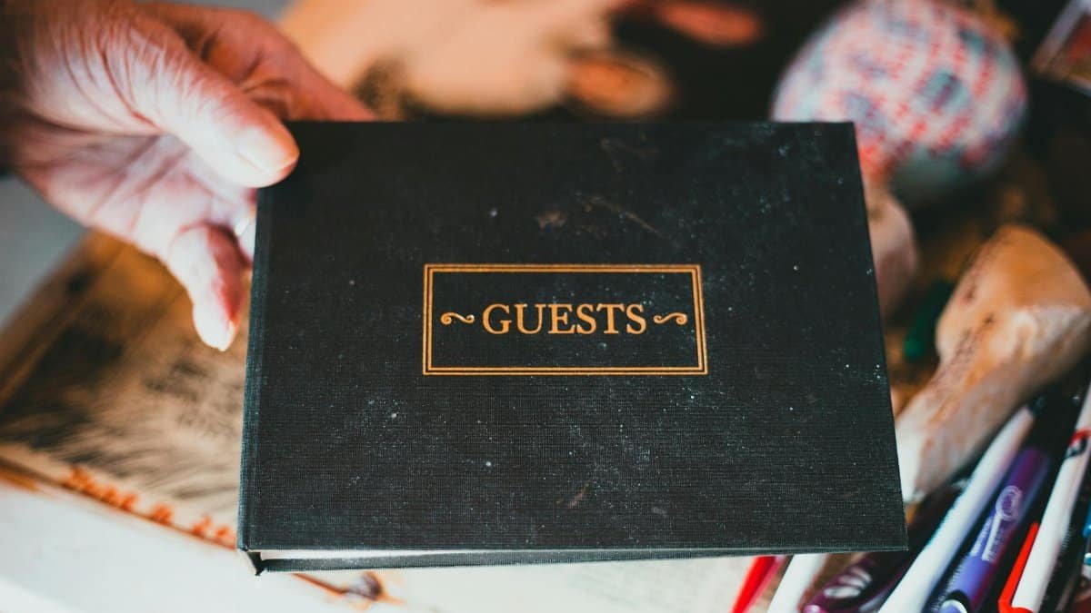 A hand holds a guest book surrounded by pens and a blurry background.