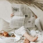 Person relaxing in bed with a newspaper, coffee, and croissant for a calm morning vibe.