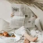 Person relaxing in bed with a newspaper, coffee, and croissant for a calm morning vibe.