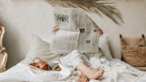 Person relaxing in bed with a newspaper, coffee, and croissant for a calm morning vibe.