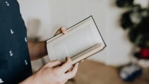 Close-up of a person holding and reading an open book indoors with a focus on hands and text.