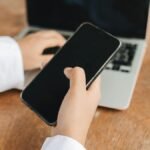Person using smartphone and laptop in a home office setup, ideal for remote work concepts.