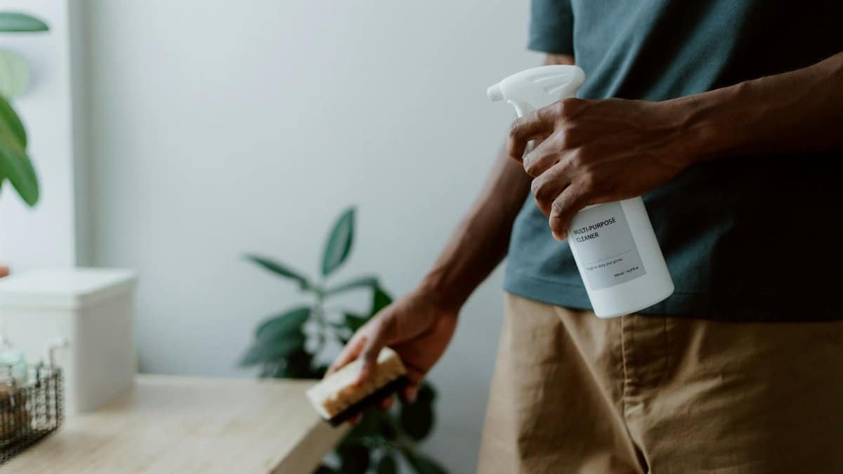 A man using a multi-purpose cleaner and sponge to clean a table indoors, emphasizing hygiene.