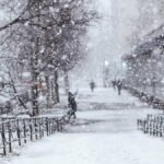 A tranquil urban scene with heavy snowfall. Bare trees and blurred figures add depth to the winter atmosphere.