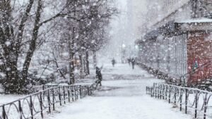 A tranquil urban scene with heavy snowfall. Bare trees and blurred figures add depth to the winter atmosphere.
