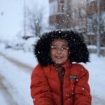A smiling child wearing a red coat enjoys a snowy day in a city street, capturing joyful winter fun.