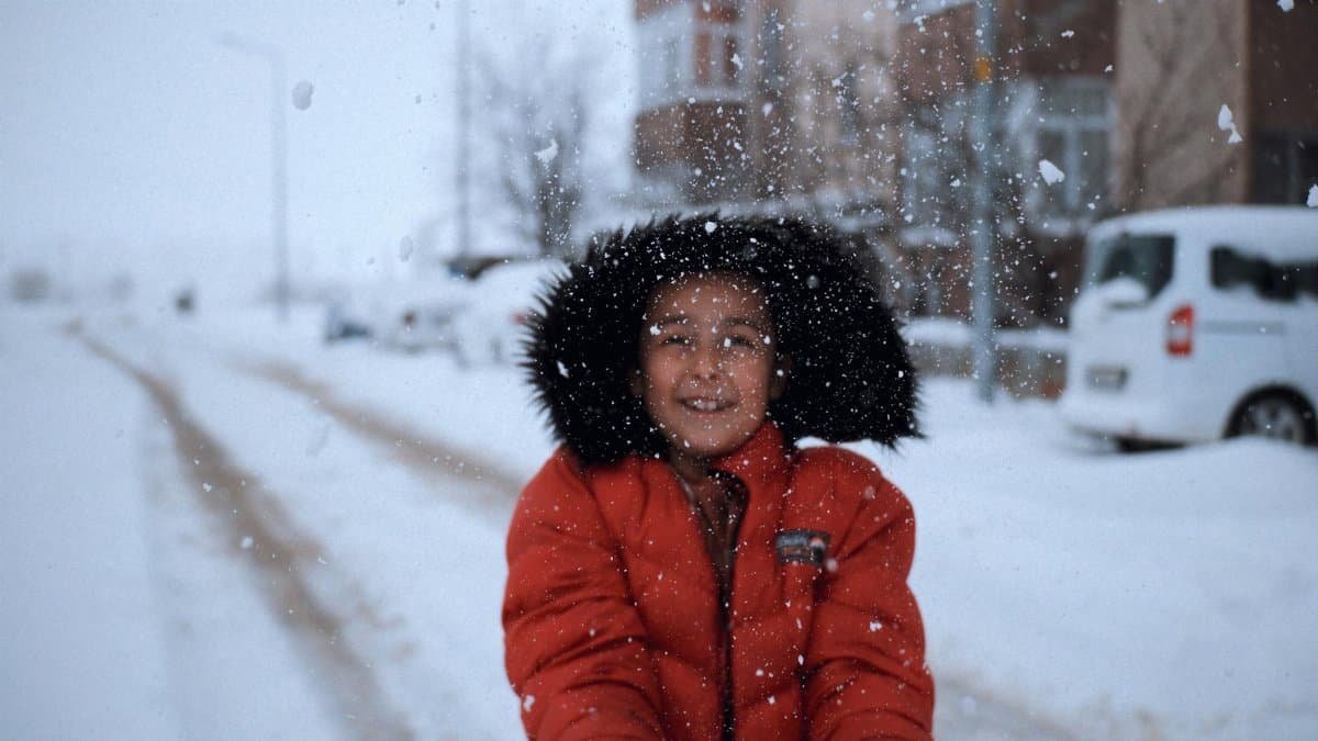 A smiling child wearing a red coat enjoys a snowy day in a city street, capturing joyful winter fun.