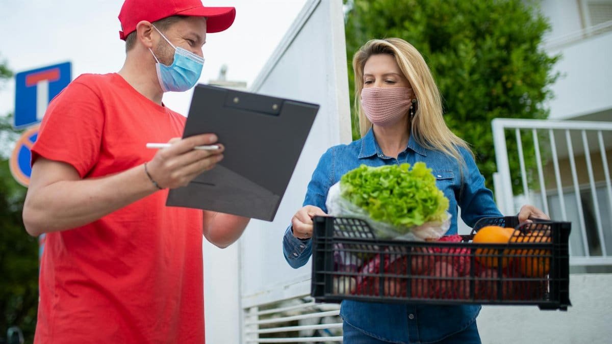 A delivery person in protective gear handing groceries to a customer at home.