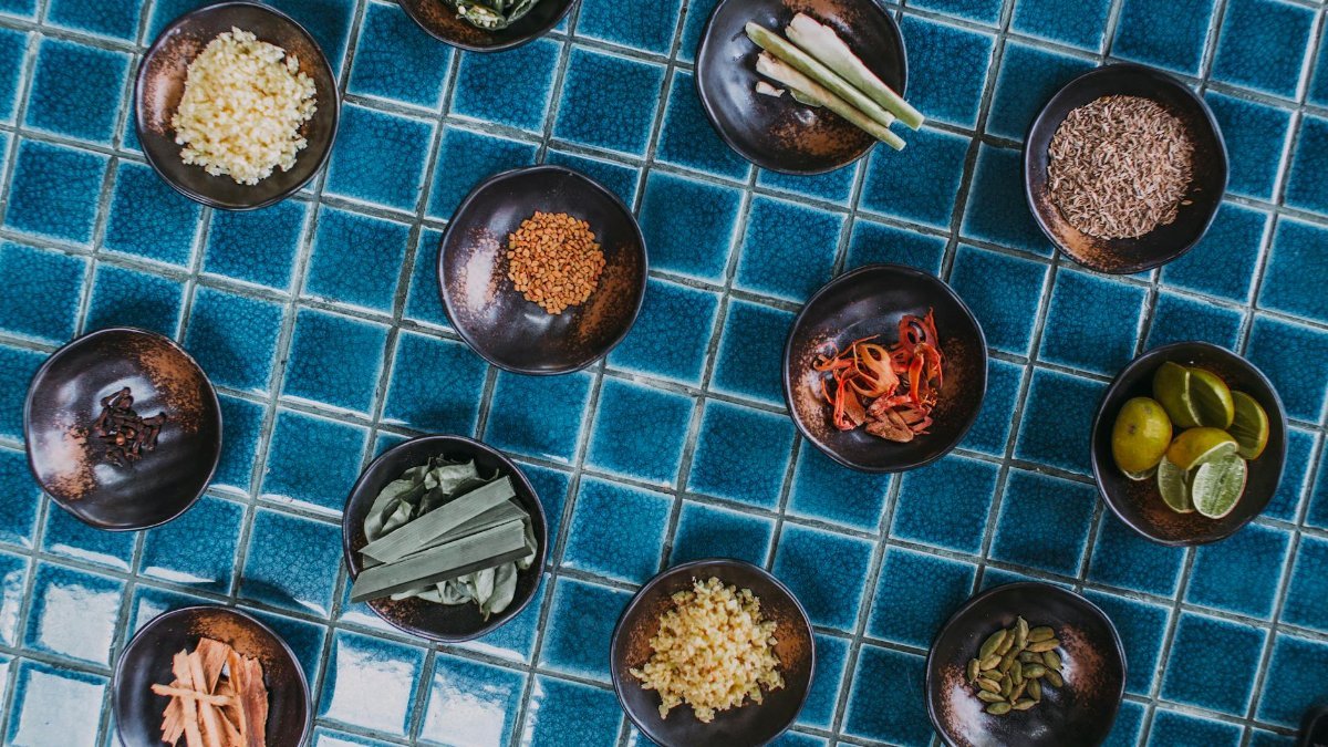 Assorted spices and ingredients in bowls arranged on blue tiles, top-down view.