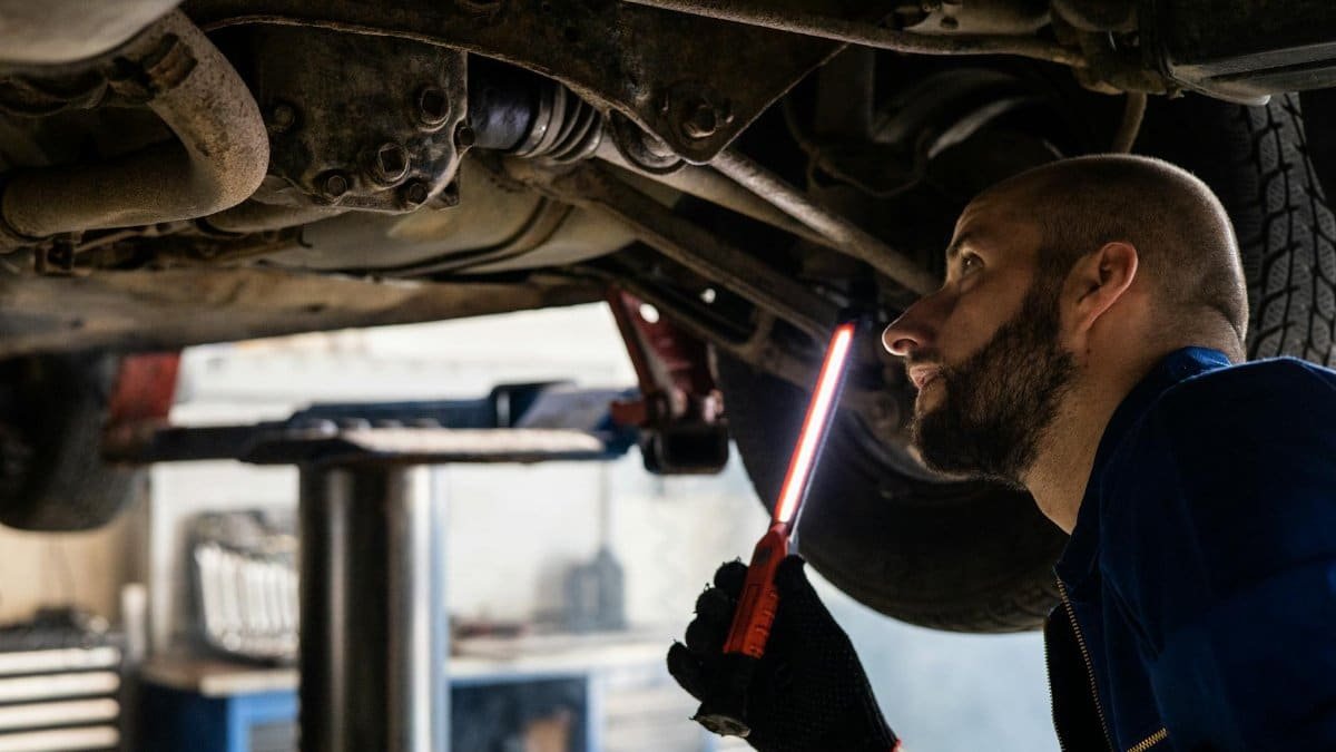Mechanic using light to inspect vehicle undercarriage in workshop for maintenance.