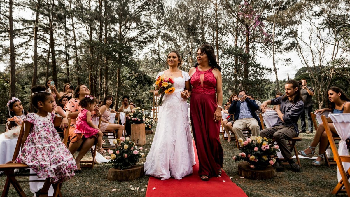 A joyful bride walking with her mother on a red carpet at an outdoor wedding ceremony surrounded by guests.
