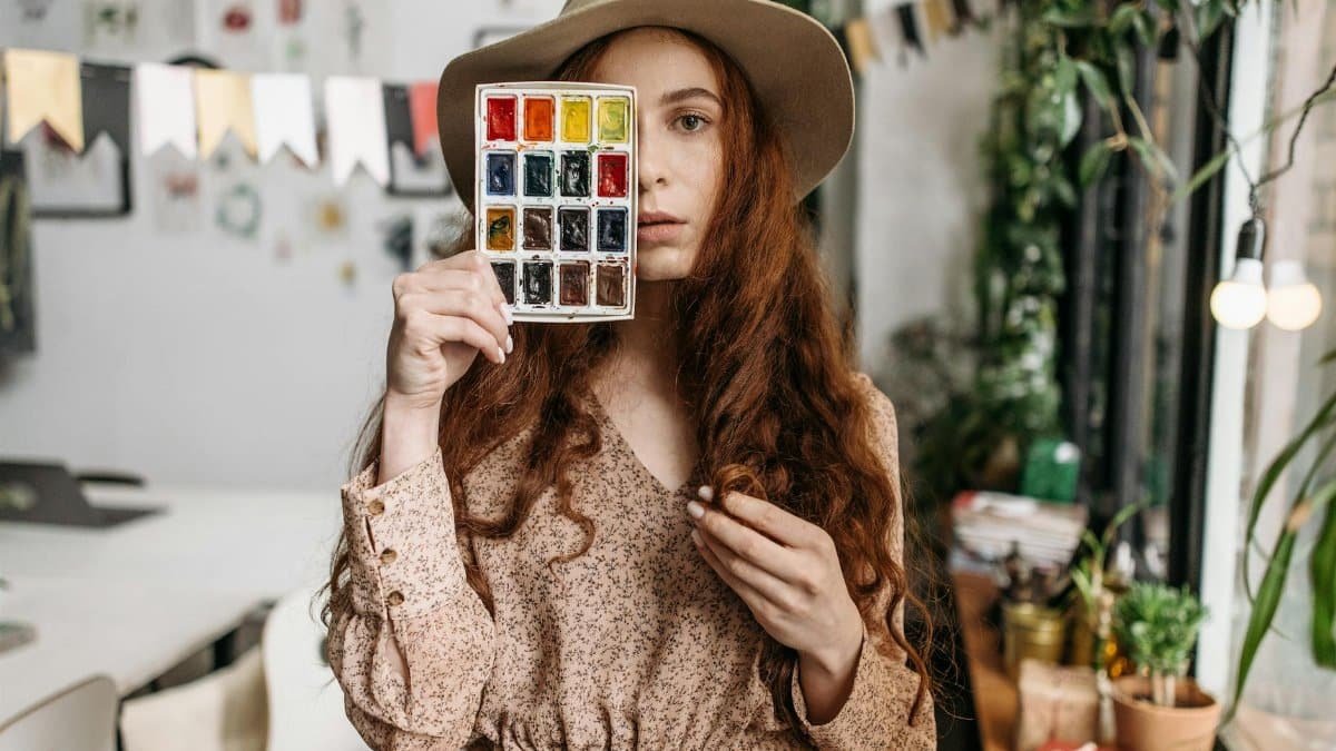 Artistic woman in a cozy studio holding a watercolor palette, surrounded by plants and creative decor.