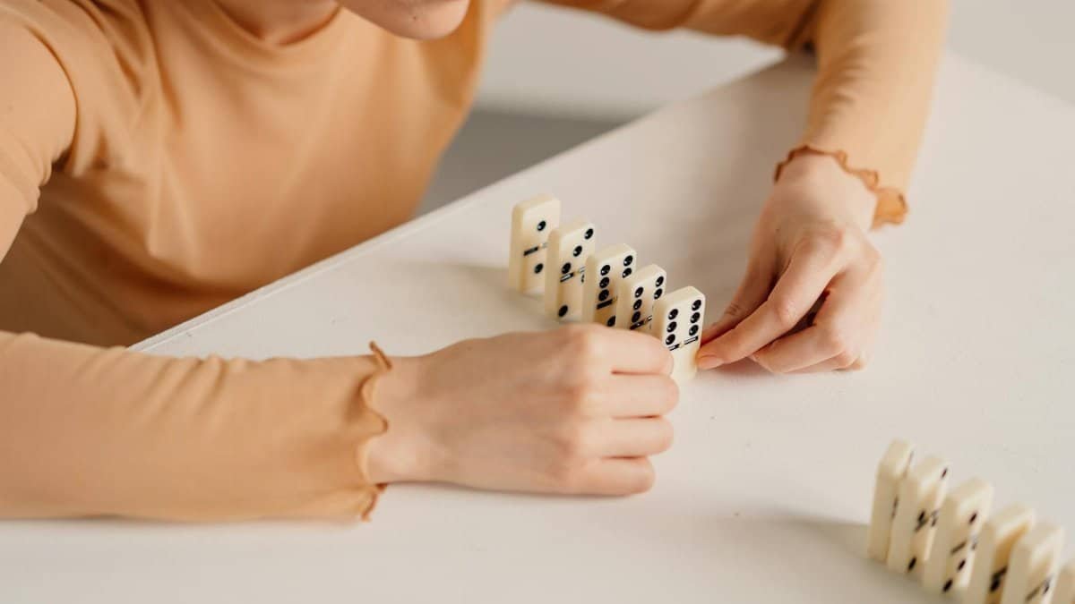A woman in beige long sleeves setting up dominoes in a line on a white table.