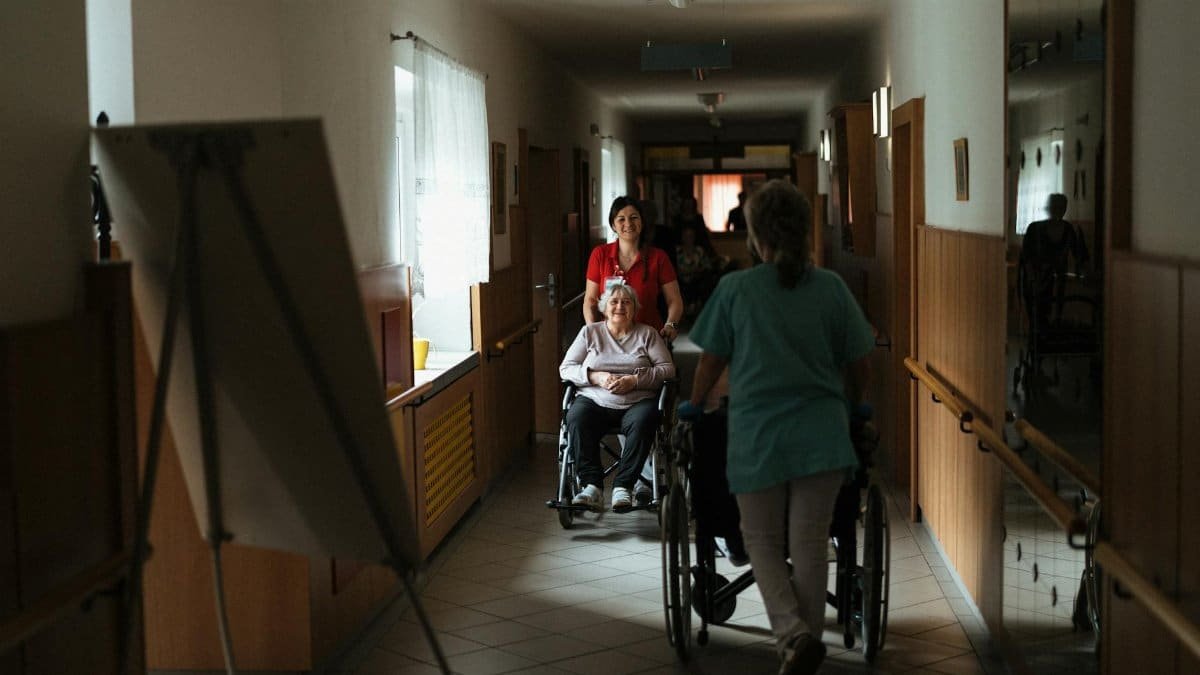 Caregiver assisting elderly woman in a wheelchair through a retirement home corridor.