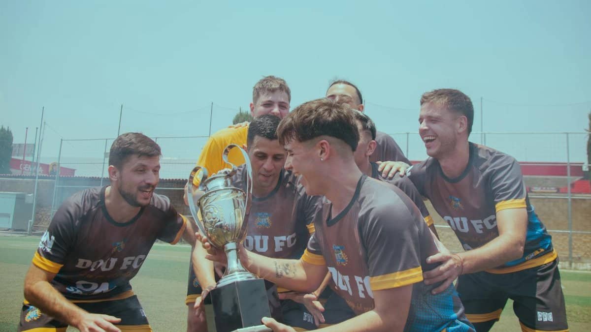 Soccer team joyfully celebrating their victory on a sunny pitch holding a trophy.