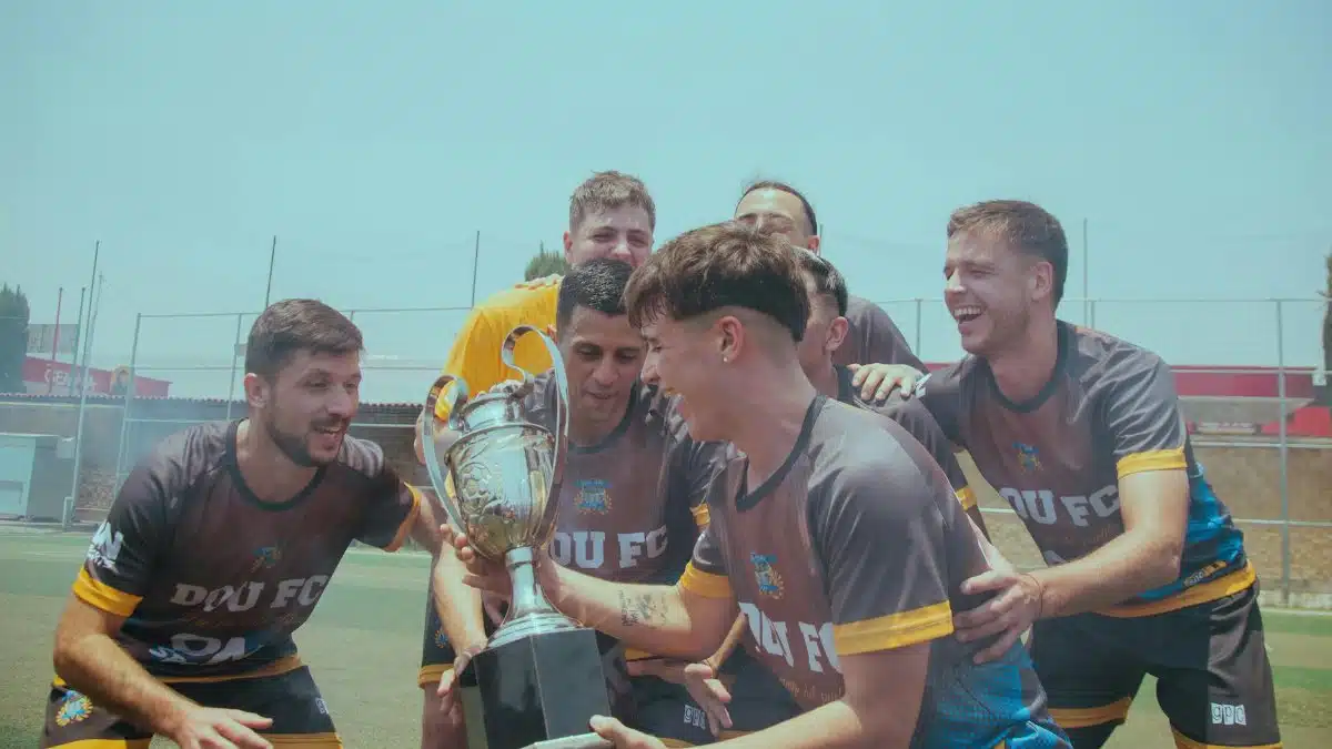 Soccer team joyfully celebrating their victory on a sunny pitch holding a trophy.