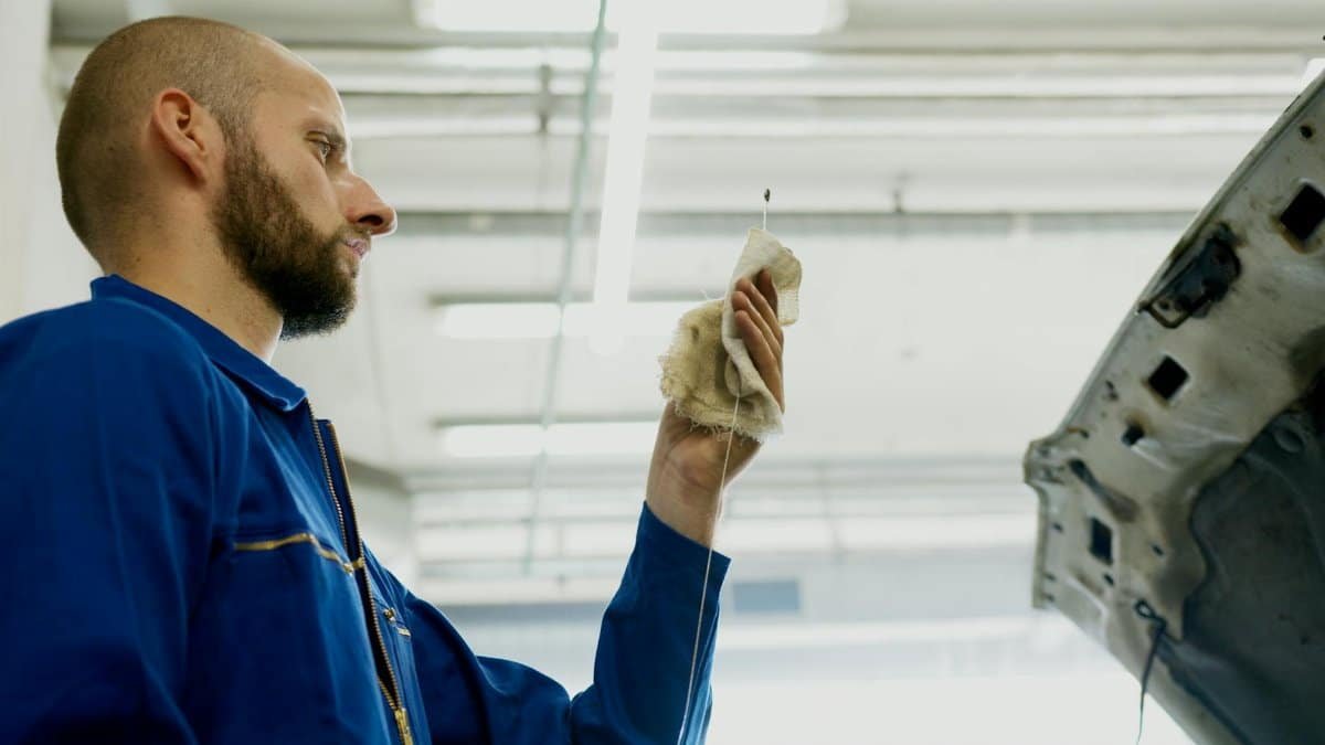 Mechanic in a blue uniform checks car oil level using a dipstick inside a garage.