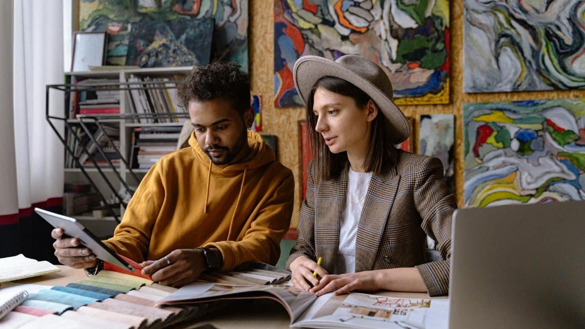 Two designers collaborate in an art-filled studio, engaging with design materials and digital devices.