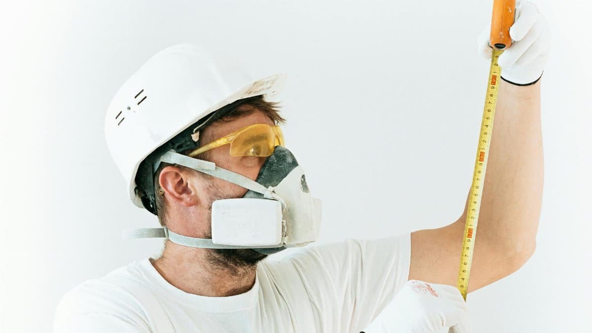Construction worker using protective gear while measuring with a tape measure indoors.