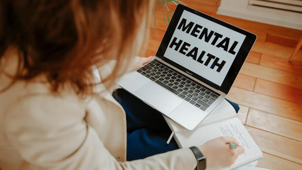 A woman participating in an online therapy session, taking notes with a laptop displaying 'Mental Health'.