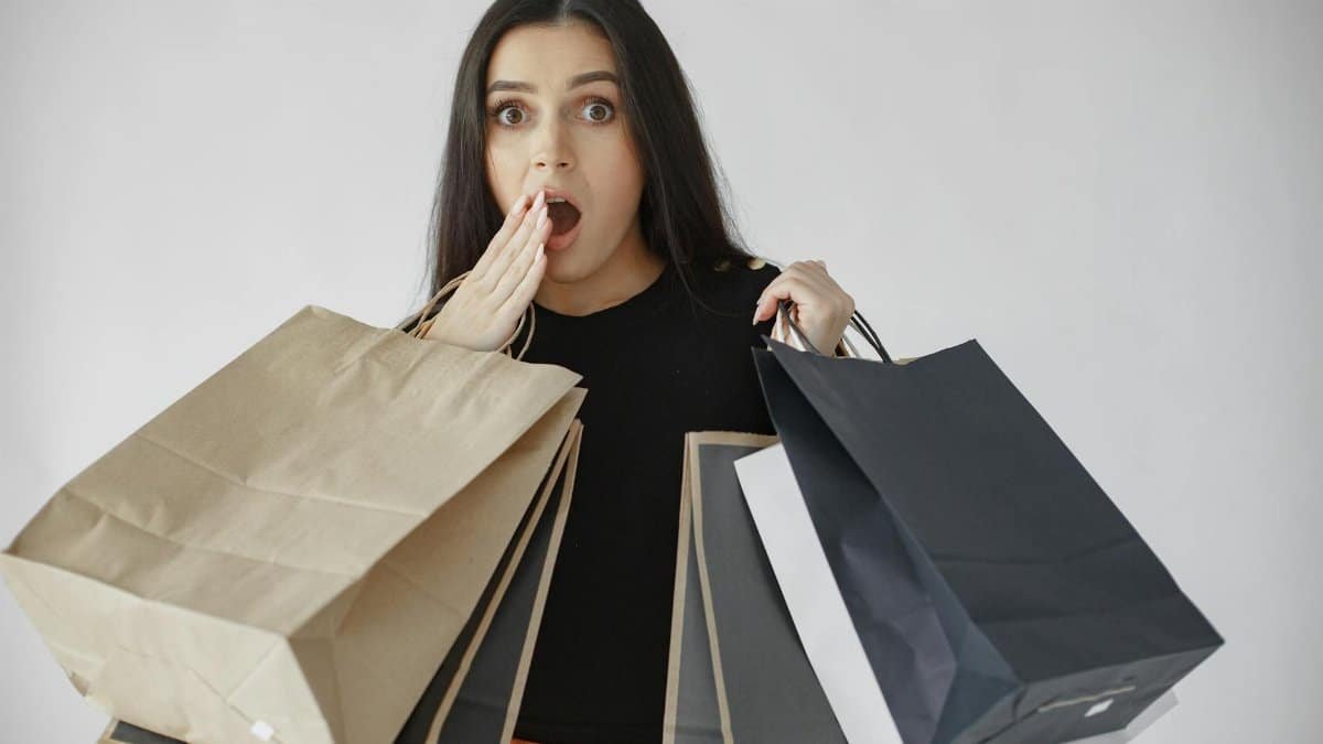 Woman shocked with shopping haul, holding multiple paper bags indoors.