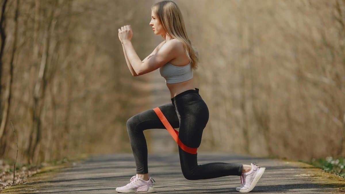 Side view of content female athlete wearing sport leggings and top performing stretching exercise with rubber resistance band while training alone in spring park
