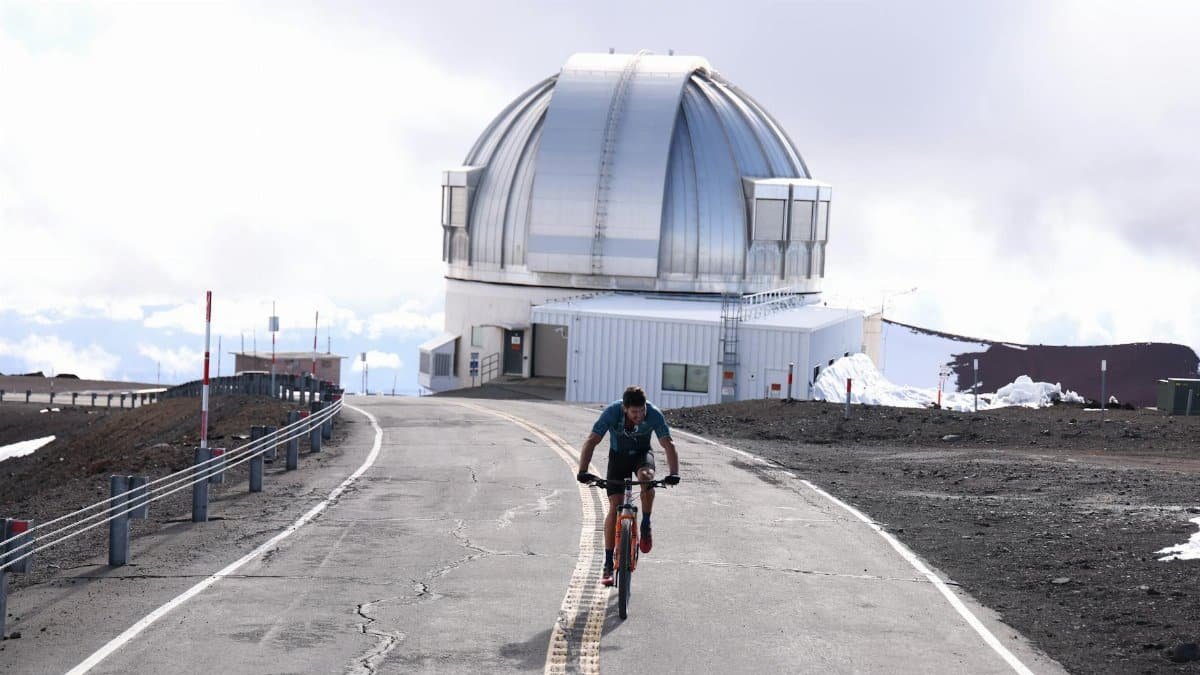 A cyclist rides towards a large observatory on a mountain road under clear skies.