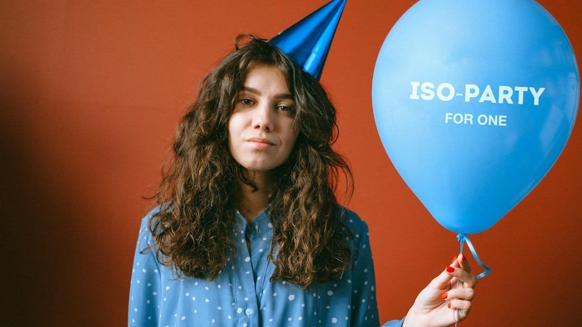 A young woman wearing a party hat holding a balloon that says 'ISO-PARTY FOR ONE.'