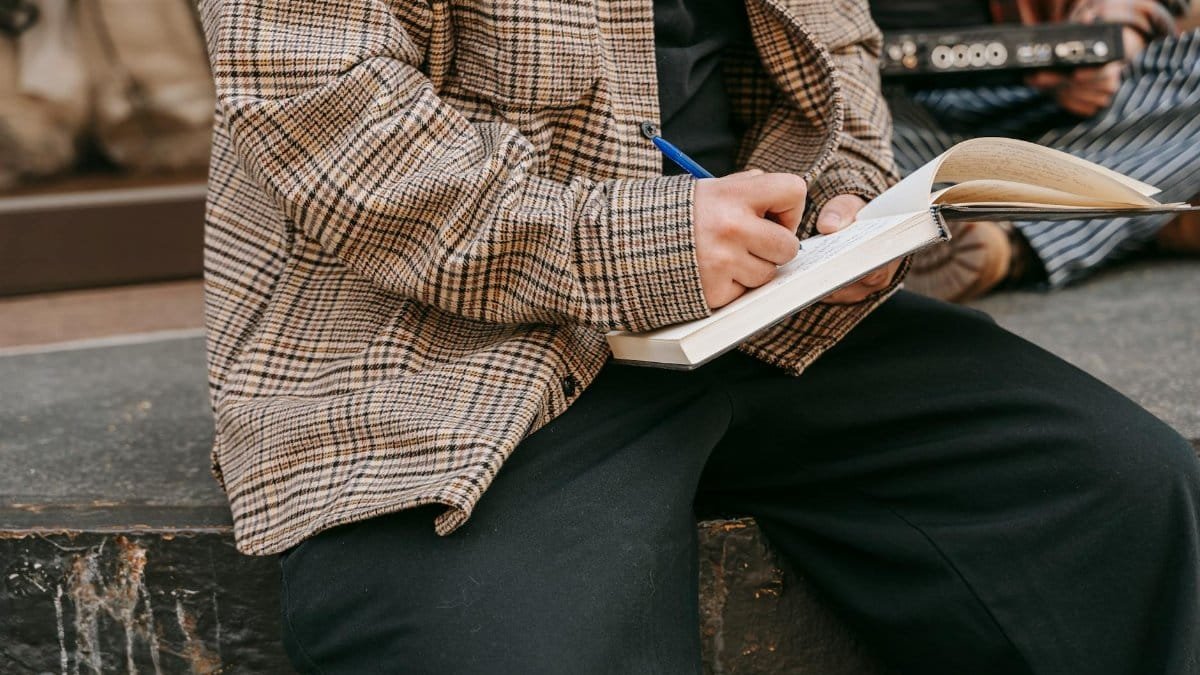 Close-up of a person writing in a notebook with a pen, sitting outdoors on a sunny day.