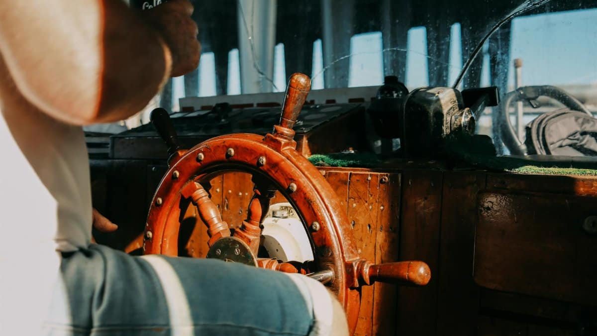A man steering a boat using a classic wooden helm in natural daylight.