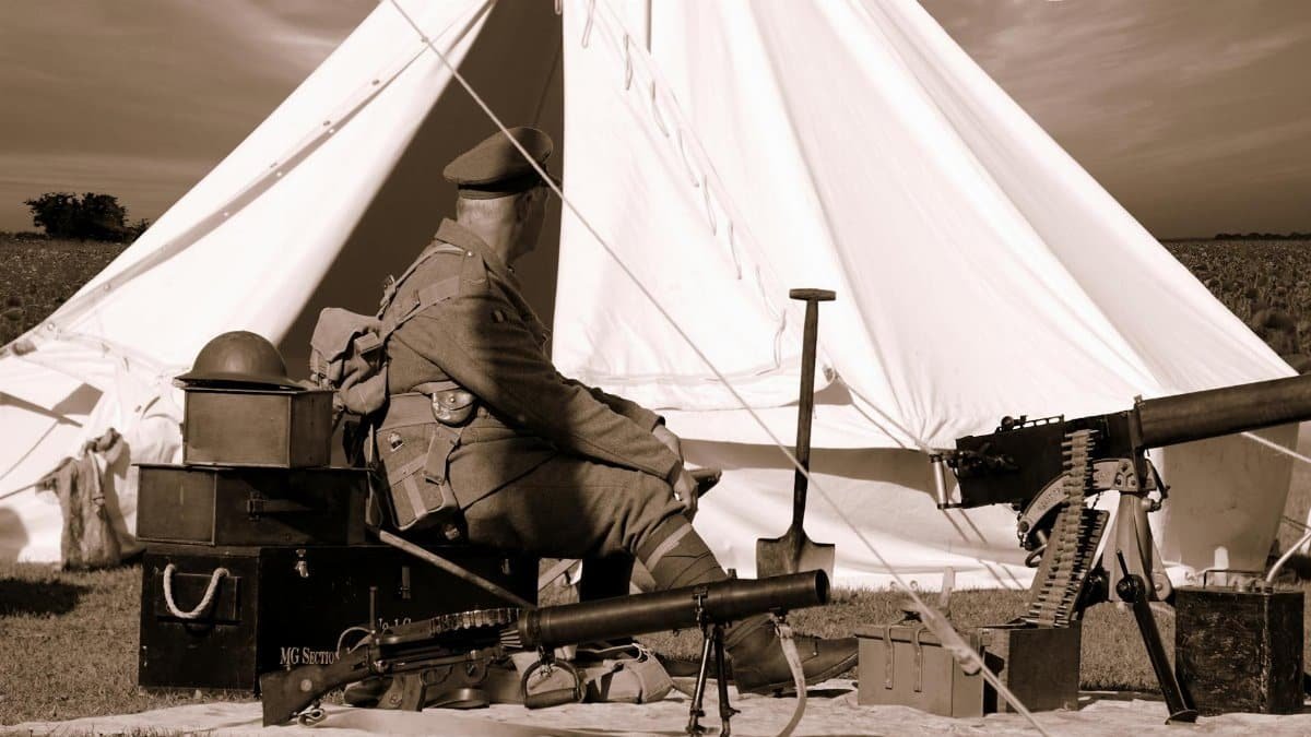 A soldier in vintage uniform sits by a tent with military equipment outdoors.