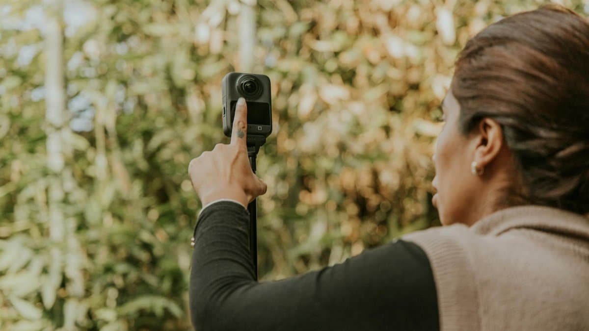 A woman adjusts a 360 camera in an outdoor setting with lush greenery, capturing a vibrant scene.