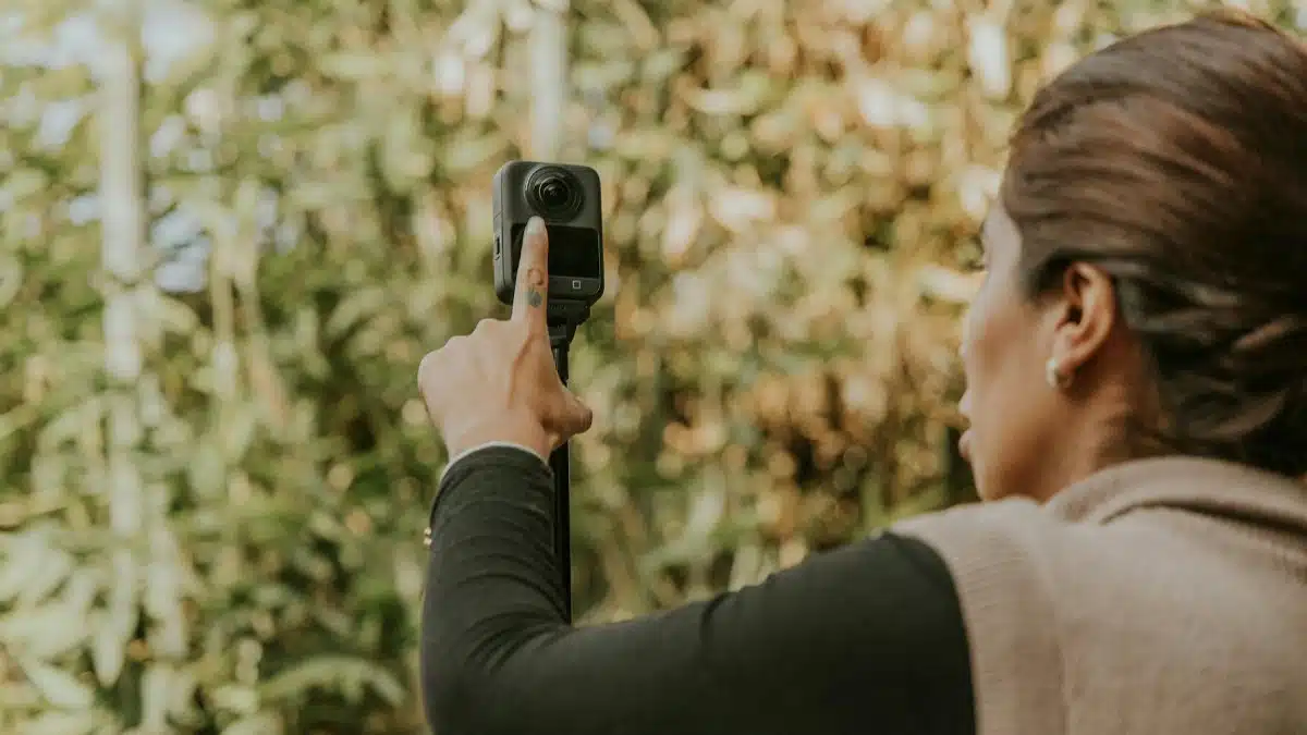 A woman adjusts a 360 camera in an outdoor setting with lush greenery, capturing a vibrant scene.