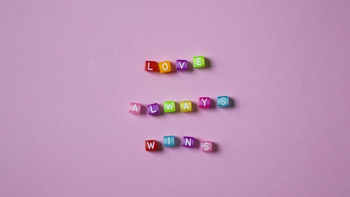 Vibrant colorful letter beads spelling 'Love Always Wins' on a soft pink background.