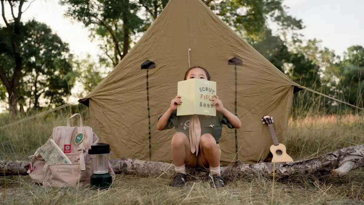 Child reading a scout field book at a campsite with a tent, ready for a camping adventure in the forest.