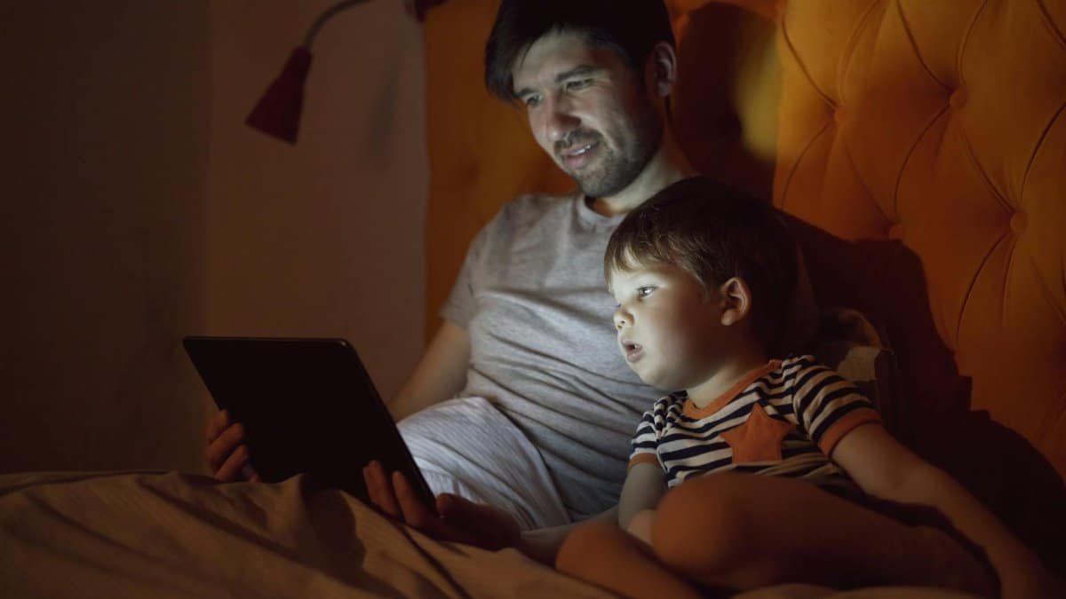 A father and young son watch a tablet together in bed at night, sharing a bonding moment.