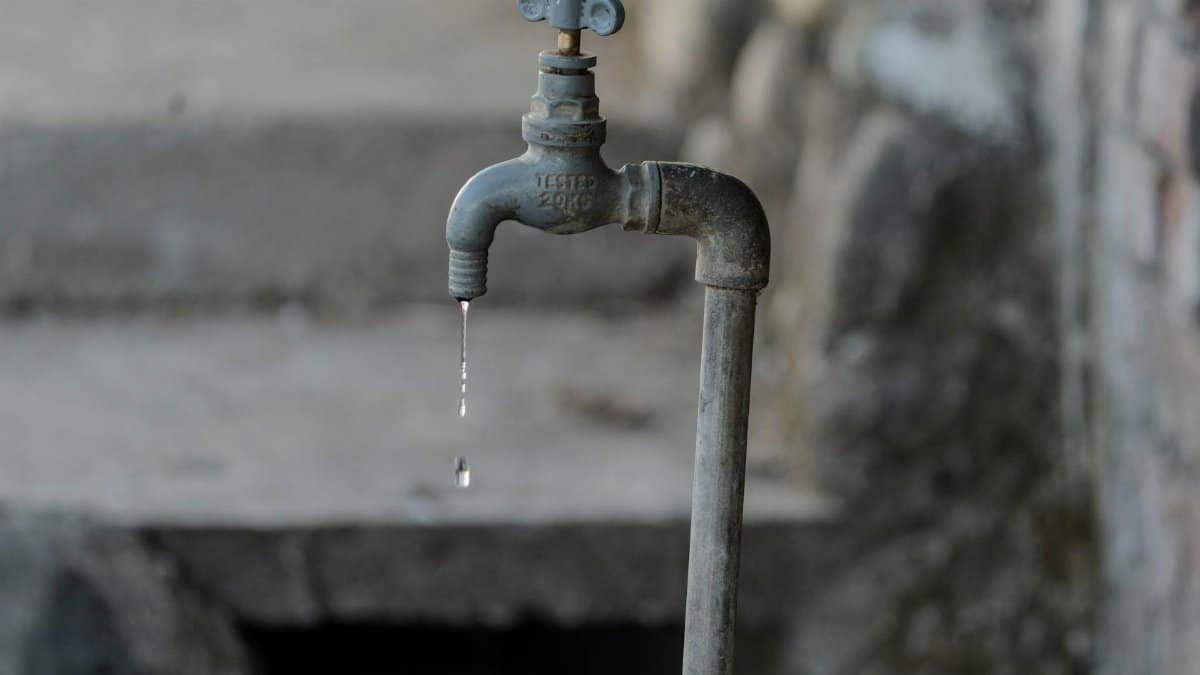 A close-up of a rustic outdoor faucet dripping water against a natural, textured background, symbolizing scarcity.