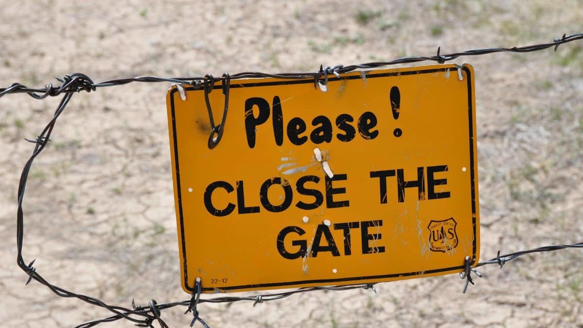 A yellow caution sign reading 'Please Close the Gate' on a barbed wire fence outdoors.