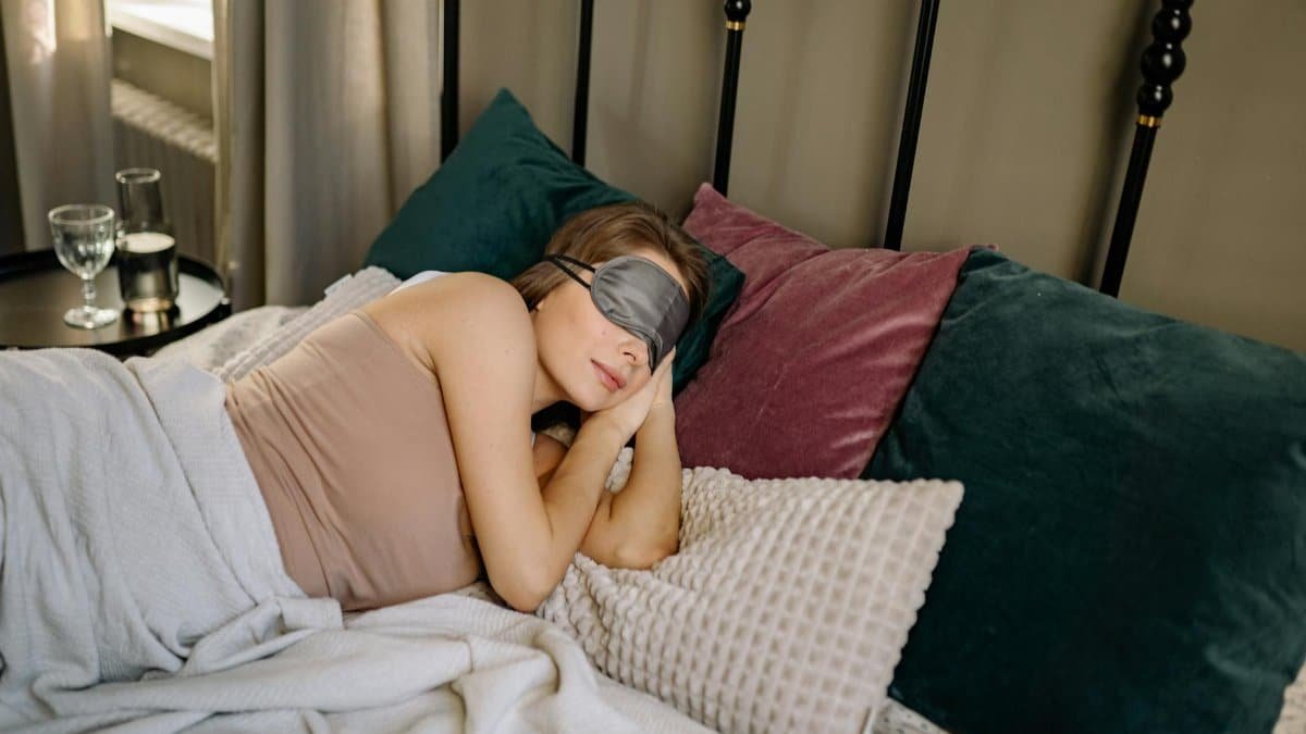Woman resting peacefully in bed with a sleep mask, surrounded by cozy pillows in an inviting bedroom ambience.