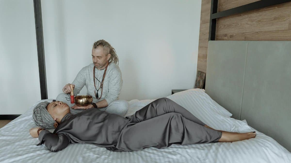 A man uses a singing bowl for sound therapy on a resting woman, promoting relaxation and spiritual healing.