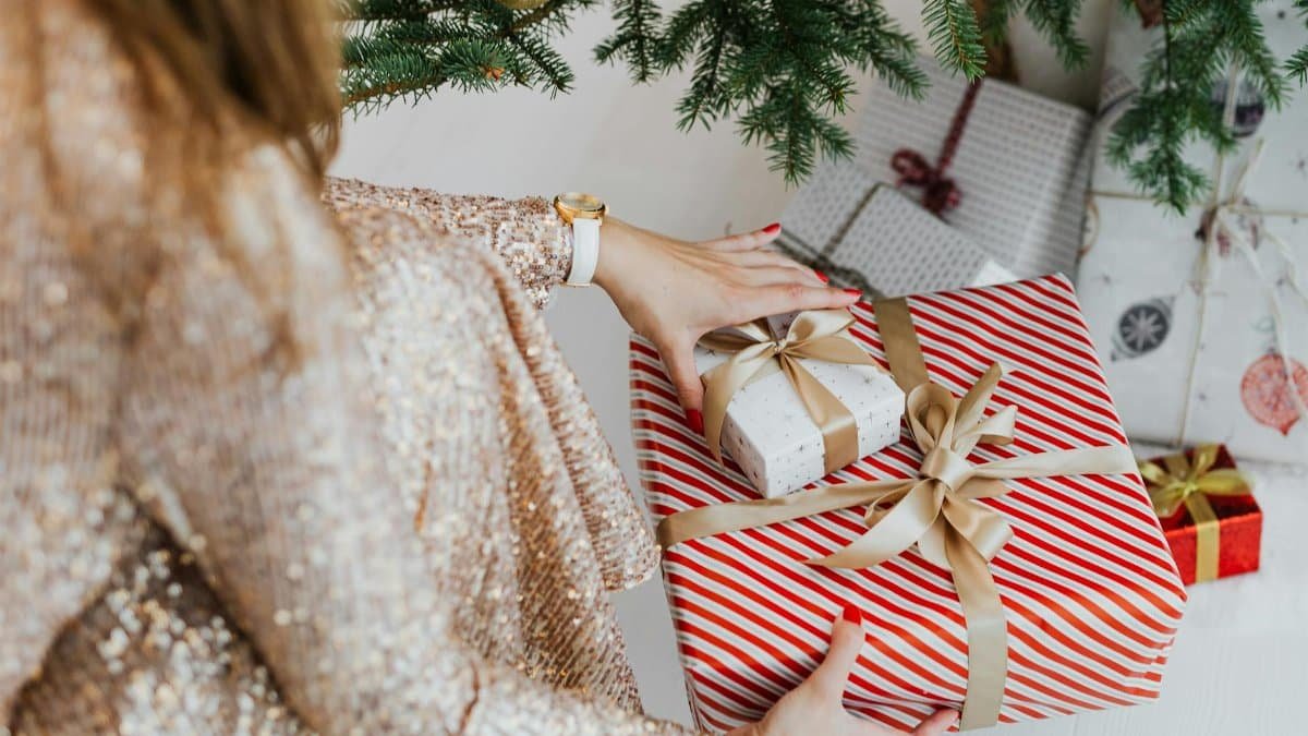 Woman crouching near Christmas tree arranging wrapped gifts at home.