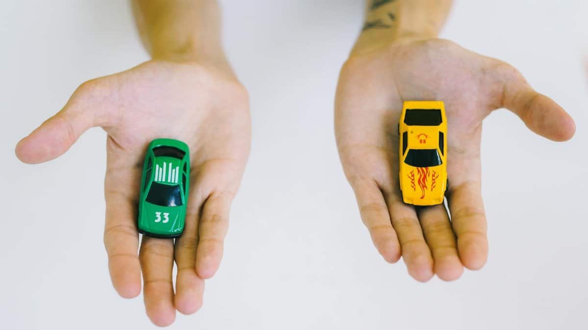 Pair of adult hands holding green and yellow toy cars on a white surface, symbolizing choice.