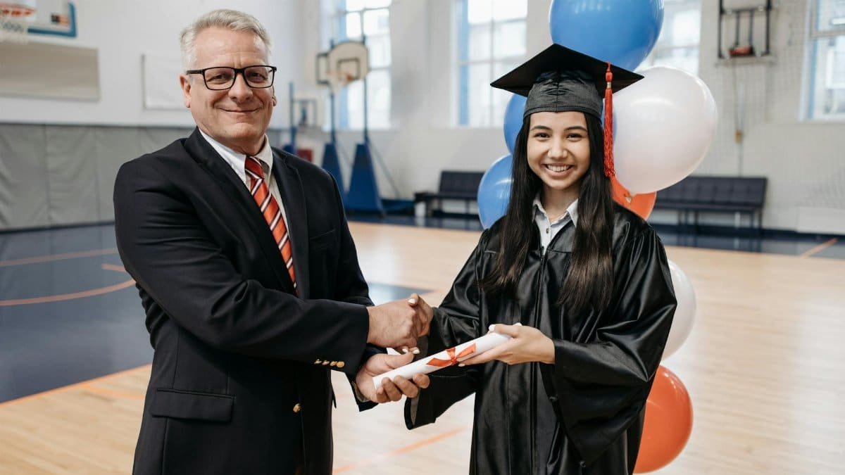 A proud graduate receiving her diploma in a gymnasium during a ceremony, shaking hands with a school director.