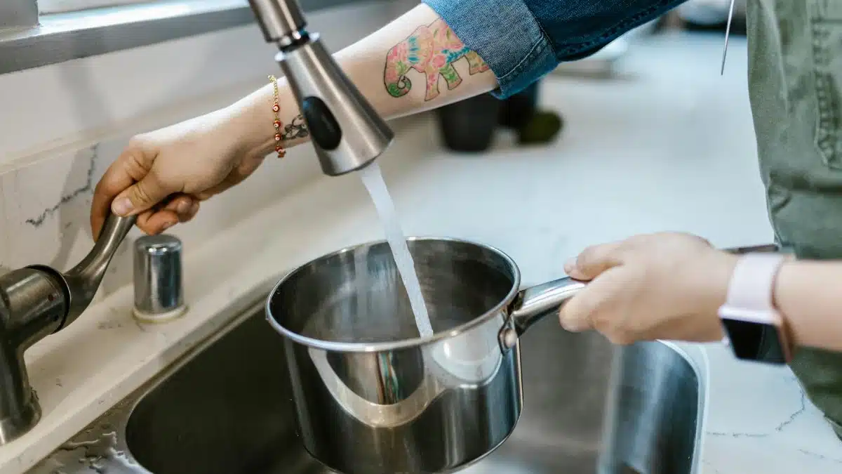 Close-up of adult hands filling a pot with tap water in a kitchen sink.